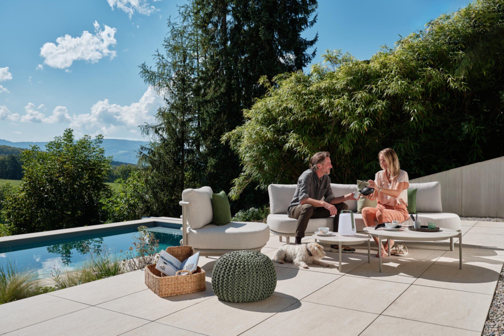 Ein Mann und eine Frau sitzen auf einer modernen Terrasse in der Nähe eines Pools und trinken gemeinsam. Sie sind von Bäumen und Bergen umgeben. Ein kleiner Hund liegt auf dem Boden, ein Korb mit Büchern und Loungemöbel stehen im Freien. Die Umgebung ist sonnig und friedlich.