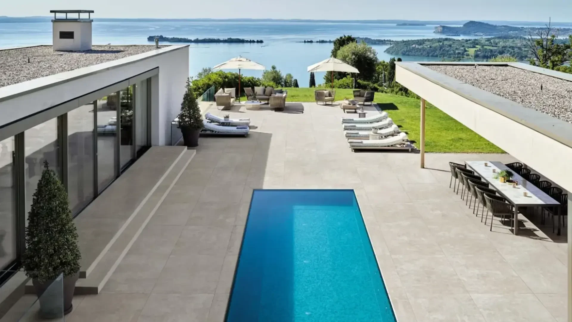 Moderne Villa mit rechteckigem Pool, Liegestühlen, Essbereich im Freien und Sitzgelegenheiten unter Sonnenschirmen, mit Blick auf einen malerischen See und eine üppige grüne Landschaft.