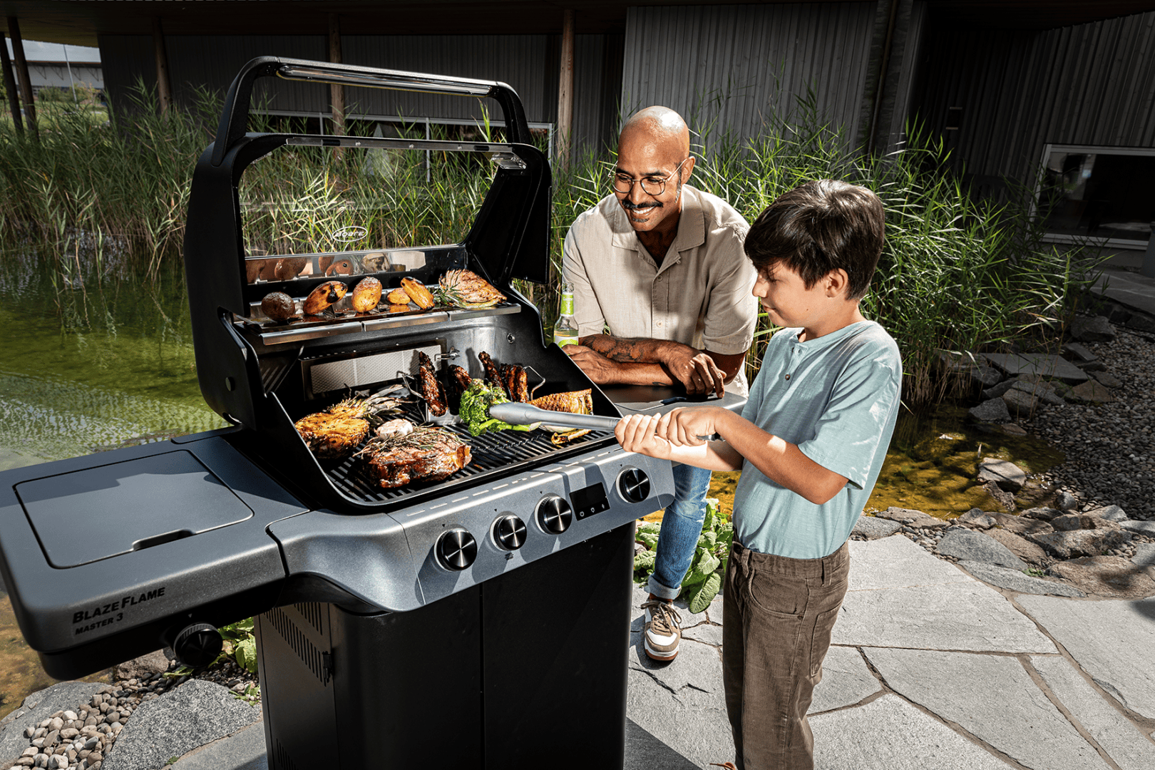 A man and a boy grill different types of meat and vegetables on a black barbecue in the open air next to a pond. They smile and enjoy cooking together on a sunny day.