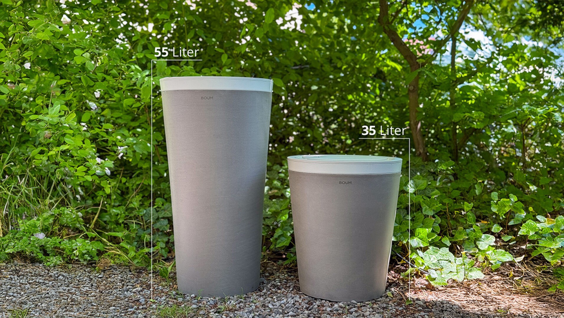 Zwei graue Mülltonnen mit weißen Deckeln stehen im Freien auf Kies, umgeben von grünen Pflanzen. Auf der größeren Tonne steht 55 Liter, auf der kleineren 35 Liter.