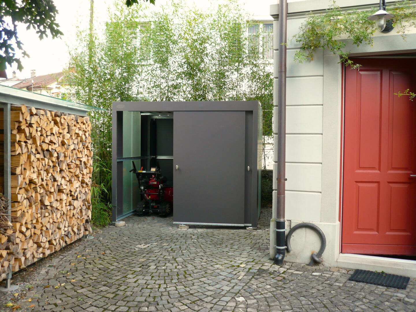 Ein moderner grauer Schuppen mit einer teilweise geöffneten Schiebetür steht auf einem Kopfsteinpflasterweg, in der Nähe eines aufgeschichteten Holzstapels und eines hellen Gebäudes mit einer roten Tür. Im Hintergrund ist etwas Grünzeug zu sehen.