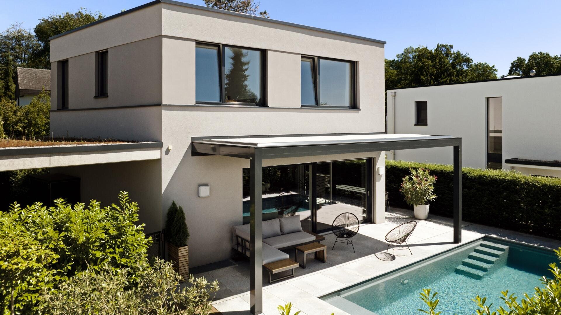 Modern two-story house with large windows, a covered terrace with outdoor seating and a swimming pool, surrounded by neatly trimmed hedges and green plants under a clear sky.