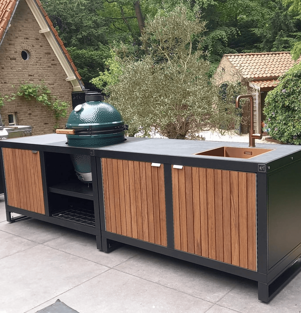 Außenküche mit einer Insel aus Holz und Metall, mit einem grünen Keramikgrill, einem Kupferwaschbecken mit hohem Wasserhahn und Aufbewahrungsschränken, auf einer Terrasse mit Bäumen und Backsteinbauten im Hintergrund.