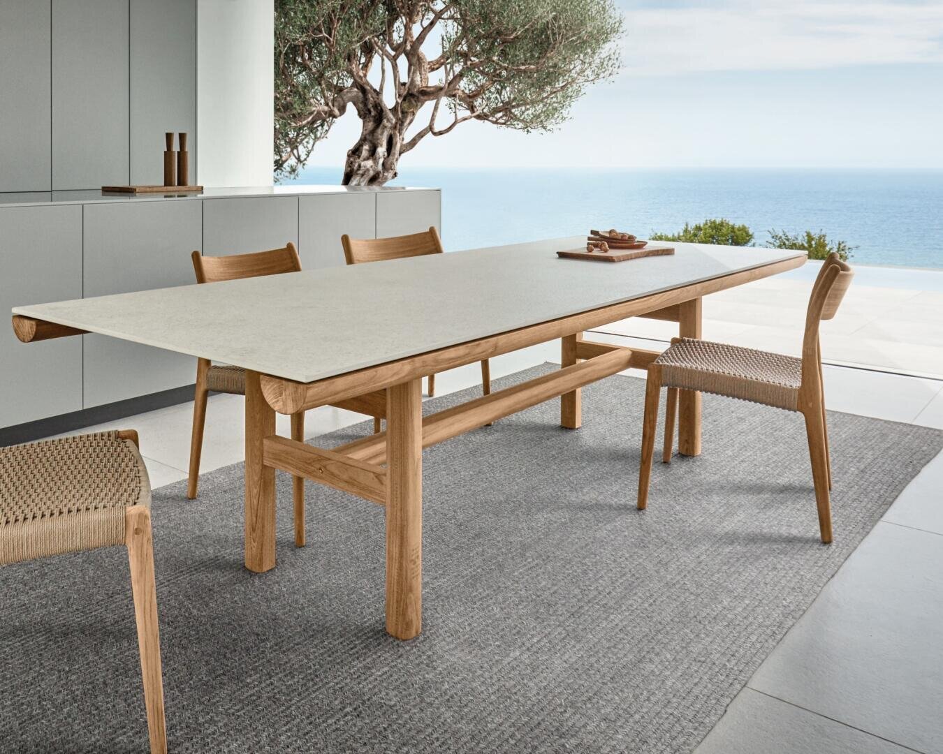 Ein moderner Essbereich im Freien mit einem langen Holztisch, vier geflochtenen Holzstühlen und einem grauen Teppich auf einer Terrasse mit Blick auf das Meer und einen Baum. Die Schränke stehen im Hintergrund.