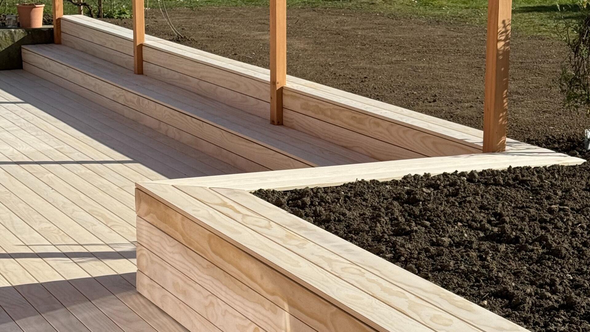Eine Nahaufnahme einer modernen Holzterrasse mit eingebauten Bänken und erhöhten Pflanzkästen, die teilweise mit Erde gefüllt sind. Die Terrasse ist aus hellem Holz, mit vertikalen Pfosten und einem Hintergrund aus nacktem Boden und Gras.
