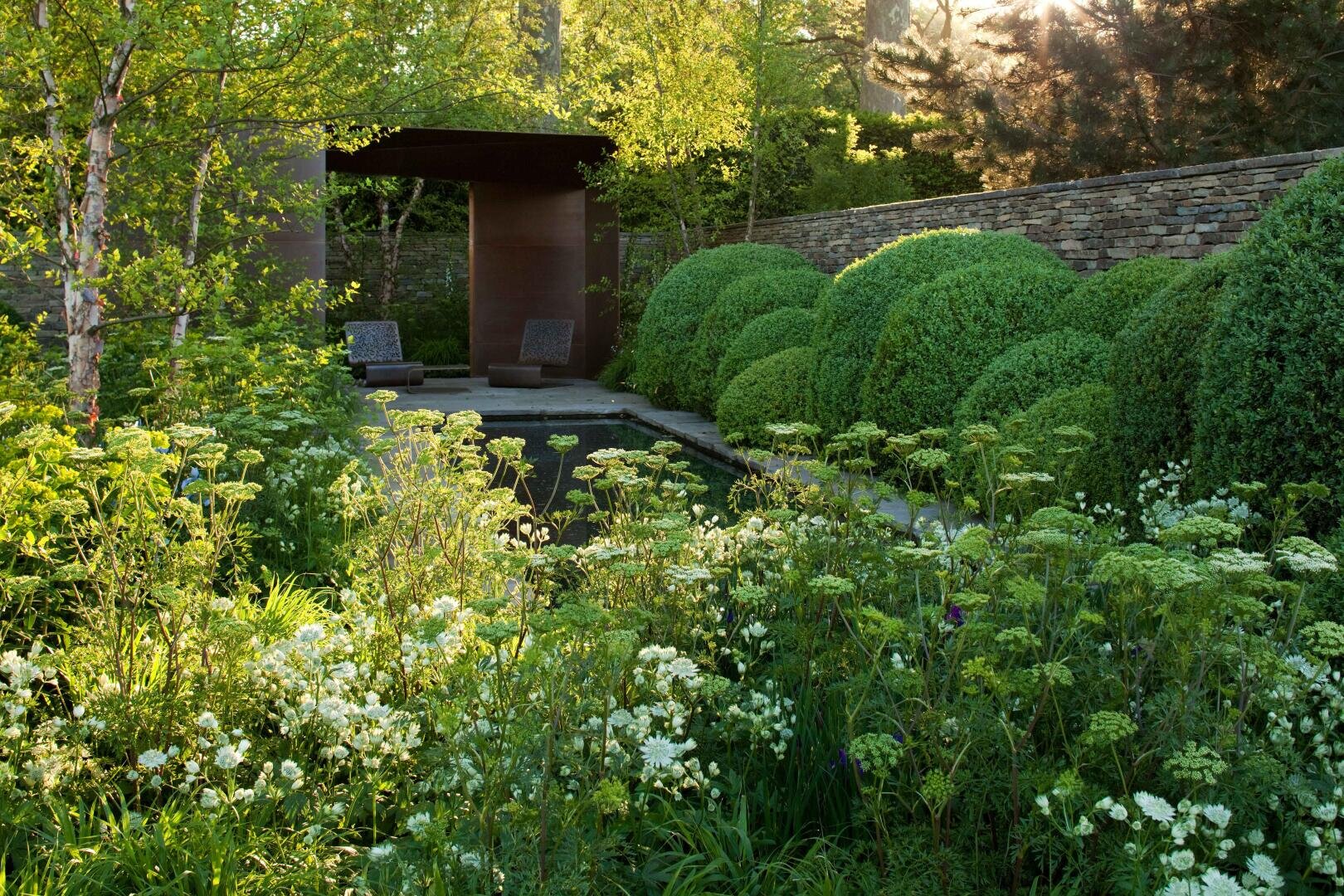 Ein üppiger Garten mit weißen Wildblumen und grünen Sträuchern umgibt einen kleinen Pool. Zwei Liegestühle stehen unter einem modernen Unterstand im Freien an einer Steinmauer. Das Sonnenlicht dringt durch die Bäume und schafft eine heitere Atmosphäre.