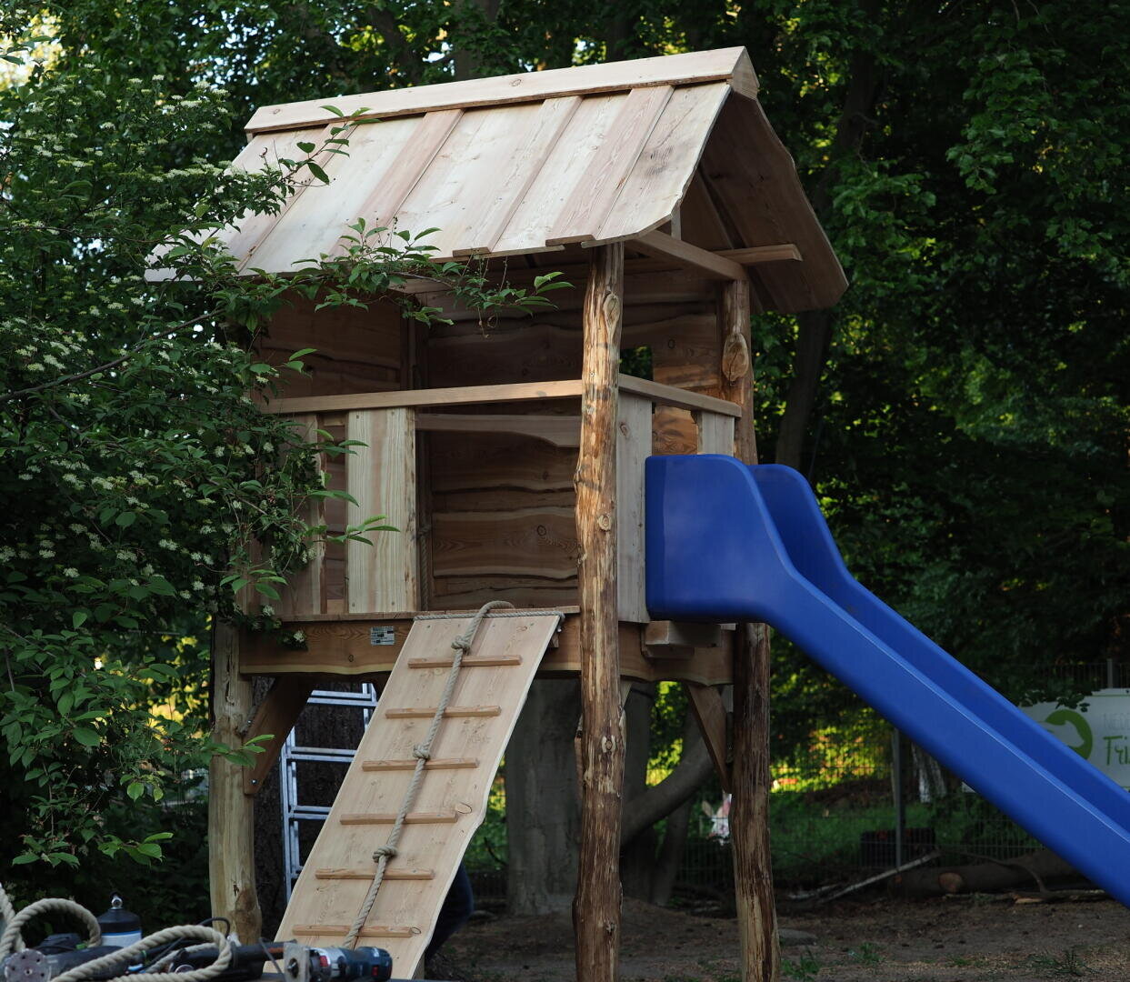 Ein hölzernes Baumhaus mit einem schrägen Dach, einer Leiter und einer blauen Plastikrutsche steht in einem schattigen, belaubten Hinterhof. In der Nähe sind verschiedene Werkzeuge und Seile zu sehen, die auf laufende Bau- oder Wartungsarbeiten hindeuten.