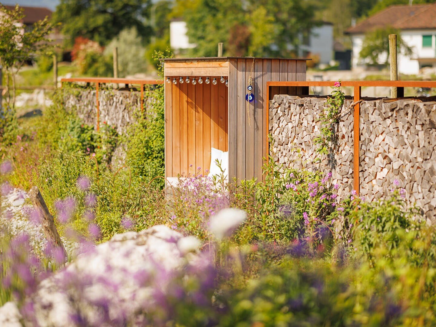 Eine hölzerne Gartendusche steht neben einem steingefüllten Zaun, umgeben von blühenden lila und weißen Blumen an einem sonnigen Tag, mit Häusern und Grün im Hintergrund.