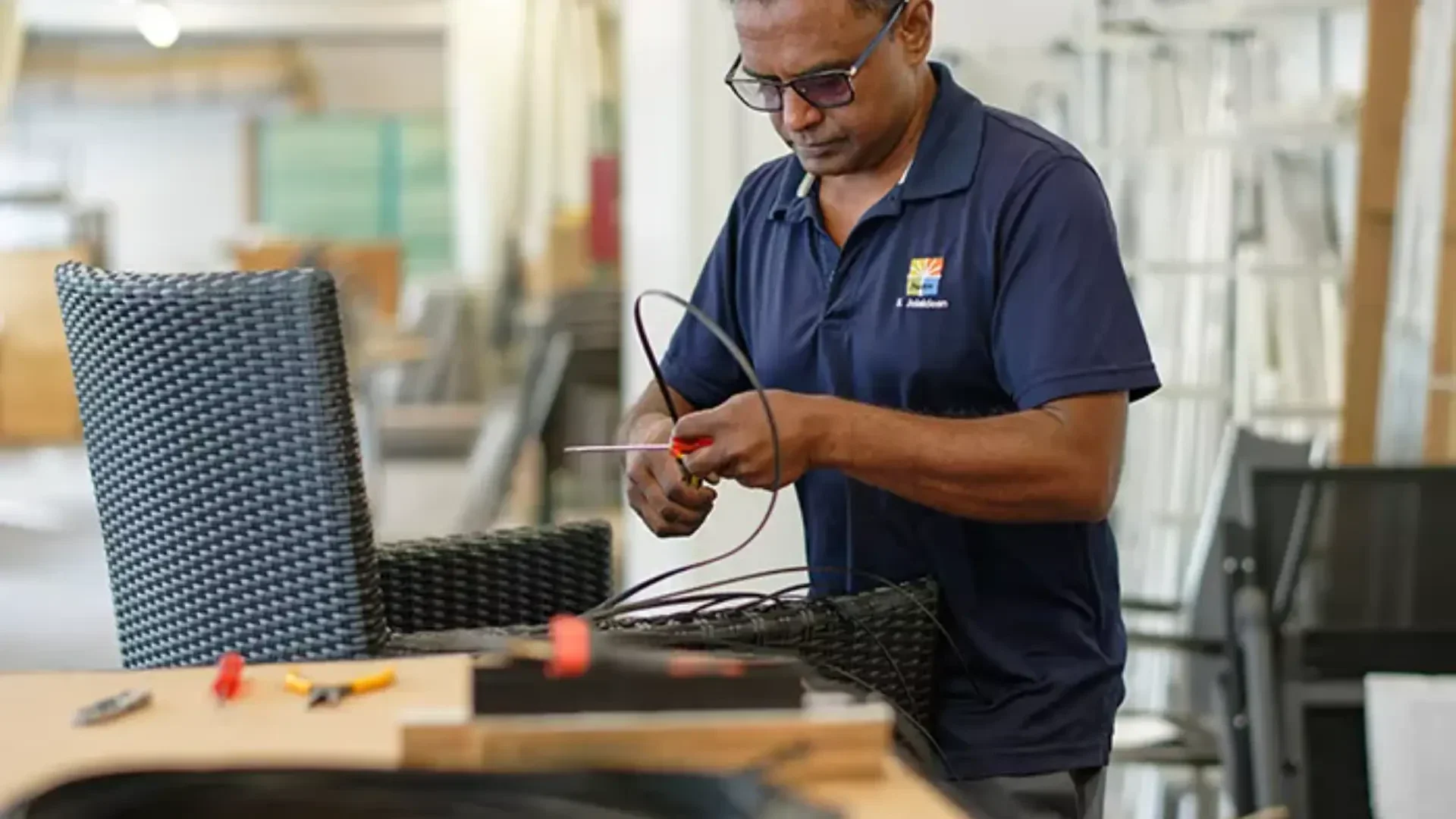 Ein Mann mit Brille und blauem Polohemd steht im Haus und arbeitet sorgfältig mit elektrischen Drähten und Werkzeugen neben einem Korbsessel und einem Tisch mit weiteren Werkzeugen.