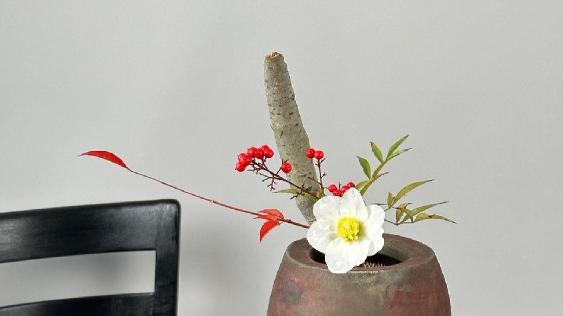 Ein minimalistisches Blumenarrangement in einer runden braunen Vase zeigt eine weiße Blume, rote Beeren, grüne Blätter und einen hohen, aufrechten Zweig vor einem schlichten grauen Hintergrund. Auf der linken Seite ist ein Teil eines schwarzen Stuhls zu sehen.