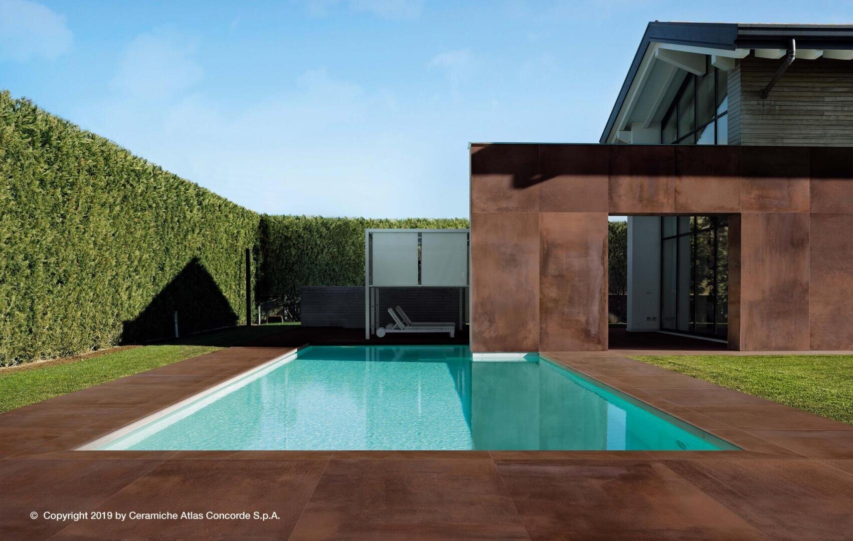 Ein modernes Haus mit großen Fenstern steht neben einem rechteckigen Swimmingpool, umgeben von braunen Fliesen und grünen Hecken unter einem klaren blauen Himmel. Am anderen Ende des Pools sind zwei Liegestühle zu sehen.