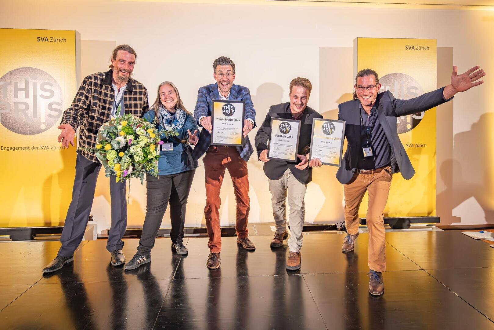 Fünf fröhliche Menschen posieren auf der Bühne, halten Auszeichnungen und einen Blumenstrauss in der Hand und feiern vor einem gelben Hintergrund, auf dem "THIS RIS" und "SVA Zürich" aufgedruckt sind.