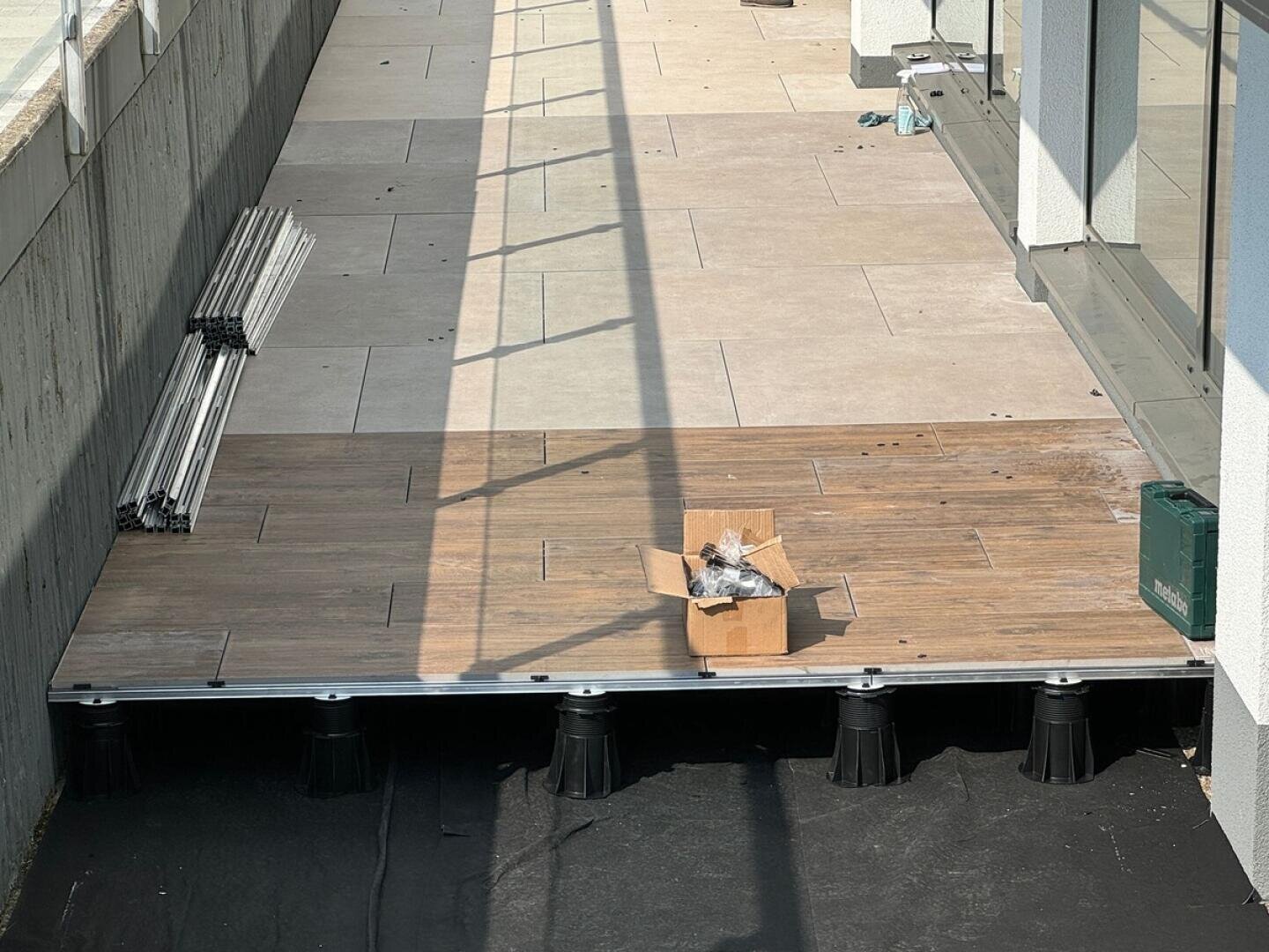 Eine teilweise fertiggestellte Außenterrasse mit großen Fliesen; in einem Bereich sind die Fliesen in Holzoptik auf schwarzen Sockeln erhöht. Auf den Holzfliesen steht eine Pappschachtel mit Materialien, und in der Nähe befinden sich gestapelte Metallstangen.