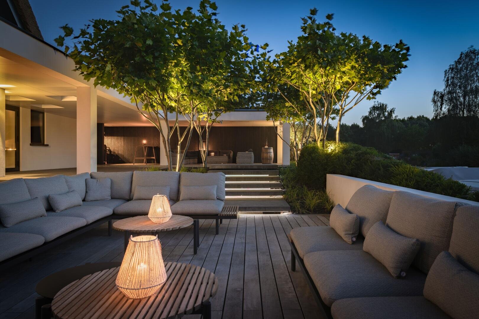 Modern outdoor terrace at dusk with cozy grey sectional sofas, glowing lanterns on a wooden table, neatly trimmed trees with overhead lighting and a softly lit house and garden in the background.