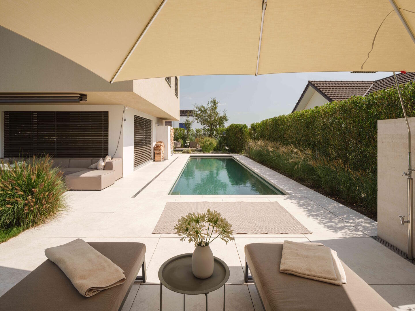 Ein moderner Hinterhof mit einem rechteckigen Pool, zwei Liegestühlen mit Handtüchern, einem kleinen Tisch mit Blumen und einem großen beigen Sonnenschirm. Eine Sitzecke und Grünflächen säumen den Poolrand.