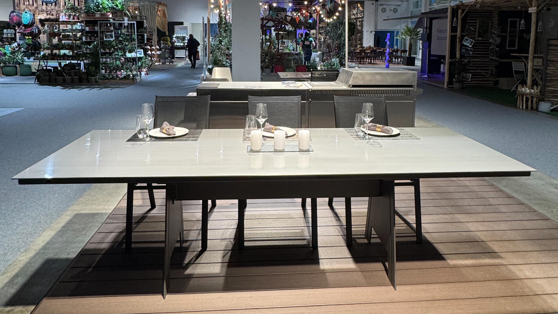 Ein moderner Esstisch mit sechs Stühlen ist mit Gläsern, Tellern und Servietten gedeckt, die in einem hell beleuchteten Raum ausgestellt sind. Die Umgebung ist mit verschiedenen Möbeln und dekorativen Pflanzen ausgestattet.