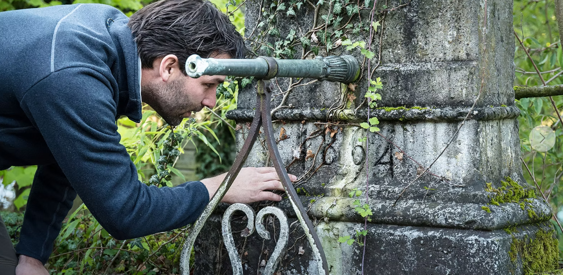 Ein Mann untersucht ein altes, mit Efeu bewachsenes Steingebäude und schaut sich die in die verwitterte Oberfläche eingeritzten Zahlen genau an.