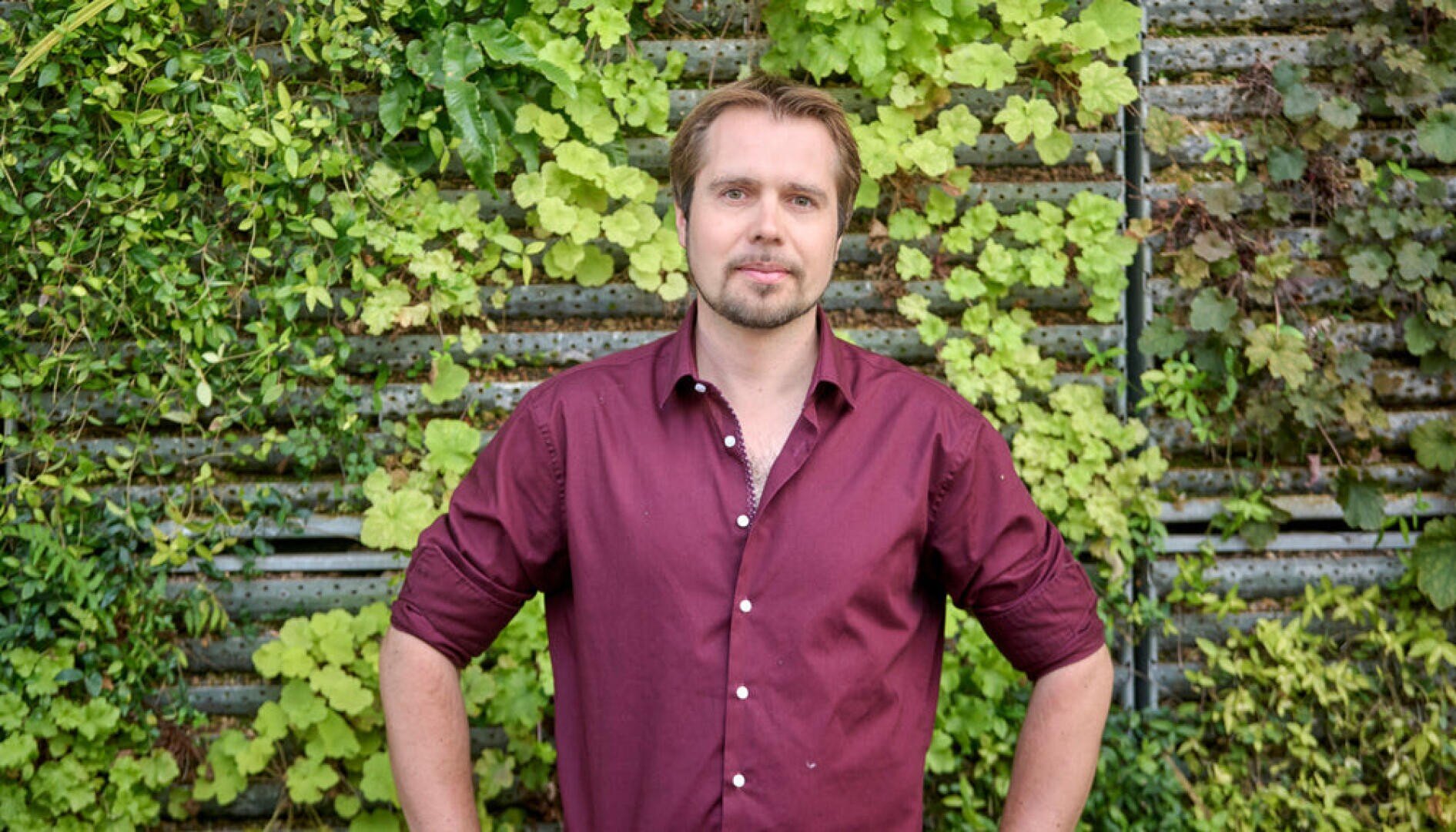 Ein Mann mit hellbraunem Haar und Bart, der ein kastanienbraunes Button-up-Hemd trägt, steht vor einer üppig begrünten Wand, die mit verschiedenen Pflanzen bewachsen ist, und blickt mit neutraler Miene und den Händen in den Hüften in die Kamera.