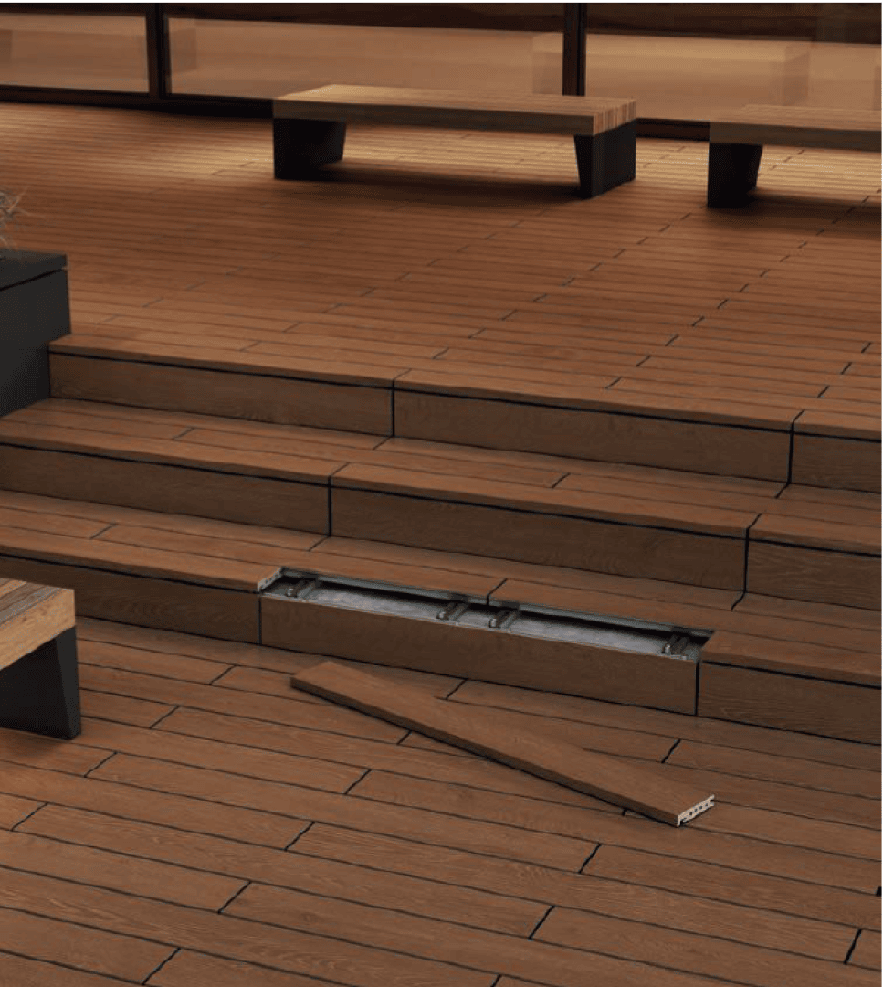 Ein Holzdeck mit Stufen ist mit einer herausnehmbaren Planke versehen, die ein Metallfach darunter offenbart. Ein freistehendes Brett liegt in der Nähe auf dem Boden; im Hintergrund sind Bänke und Pflanzgefäße zu sehen.