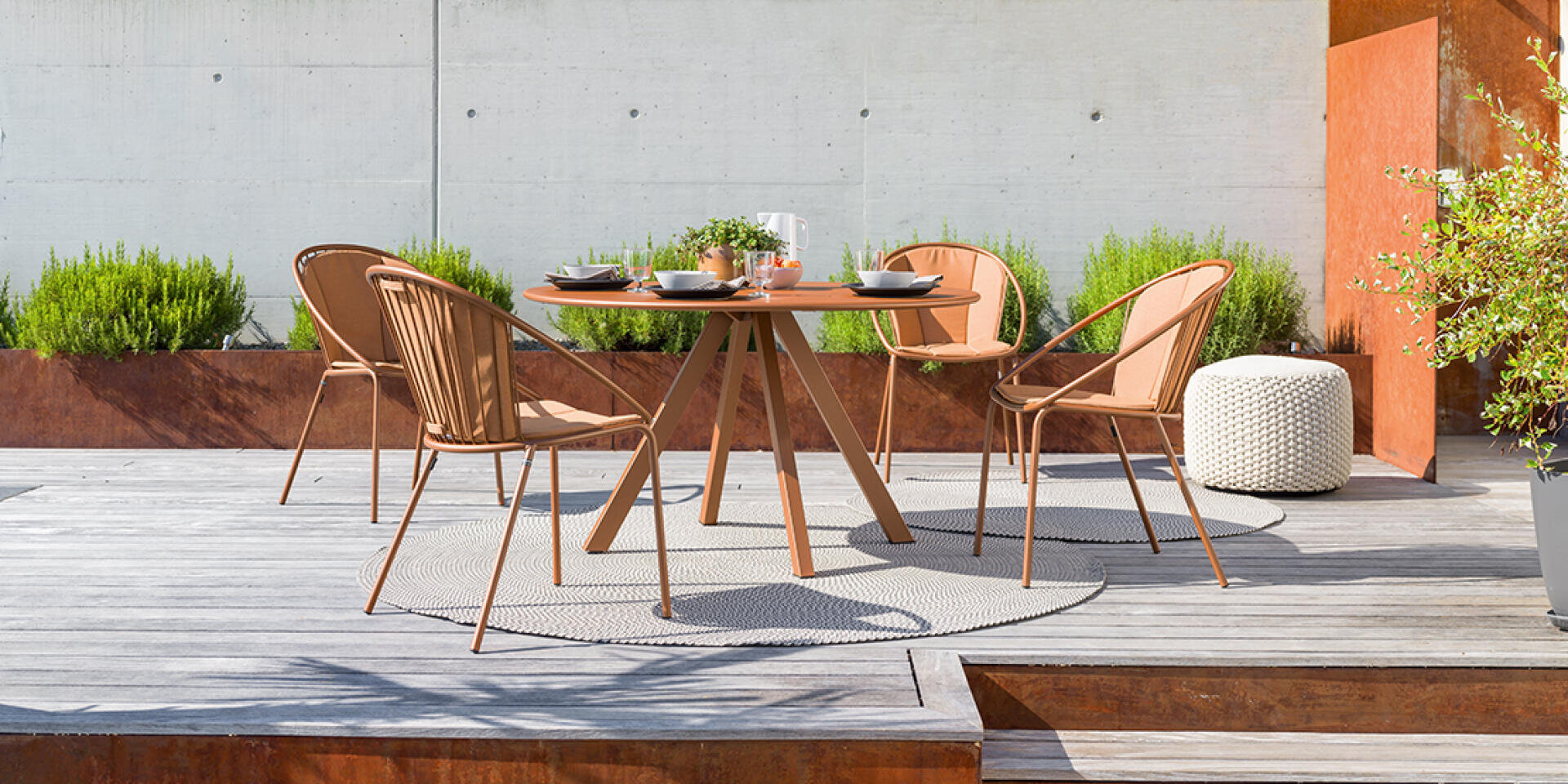 Ein runder Tisch im Freien mit vier passenden Stühlen steht auf einer Holzterrasse. Der Tisch ist mit Tellern und einer Teekanne gedeckt, umgeben von Grünpflanzen und einer Betonwand. Ein Hocker und Pflanzen schmücken den Raum.