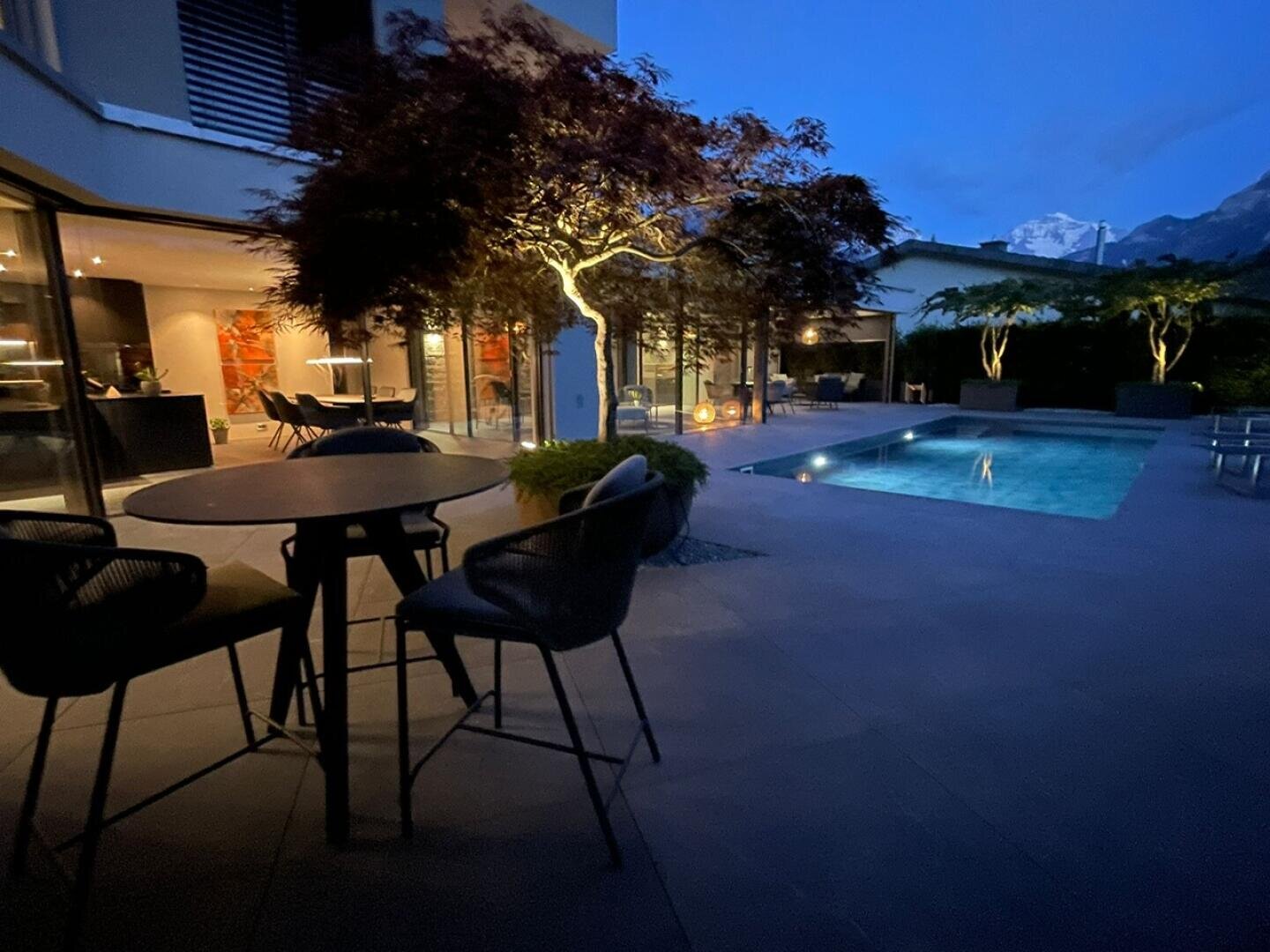 Moderner Innenhof in der Abenddämmerung mit einem beleuchteten Swimmingpool, einem runden Tisch mit Stühlen im Vordergrund, einem beleuchteten Baum und großen Fenstern, die den Blick auf einen stilvollen überdachten Essbereich freigeben. Im Hintergrund sind die Berge zu sehen.