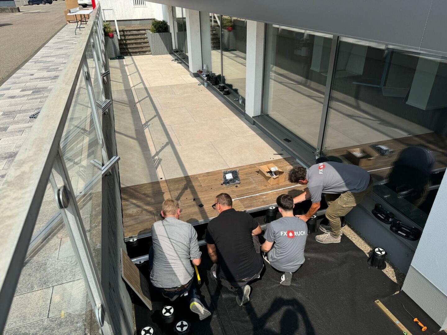 Vier Männer montieren an einem sonnigen Tag eine Terrasse vor einem Gebäude, indem sie Holzbretter und große Fliesen zusammensetzen. Werkzeuge und Baumaterialien liegen verstreut auf der Baustelle.