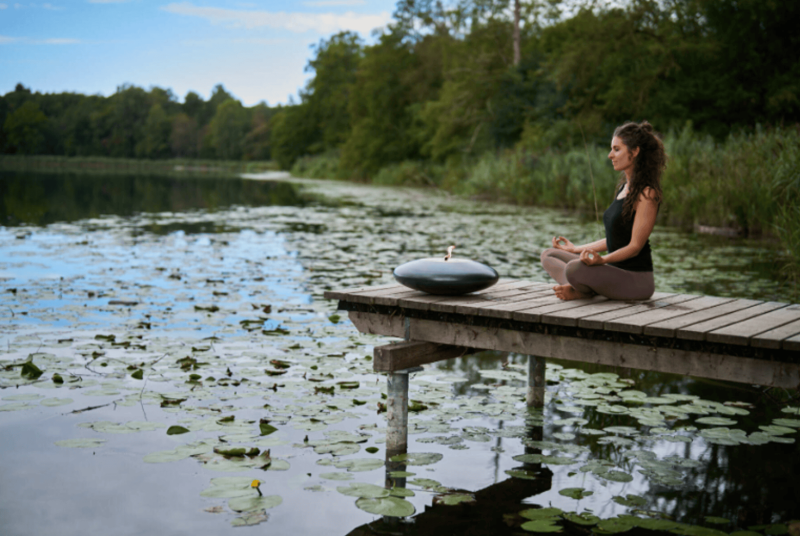 A woman sits cross-legged on a wooden dock over a lily pad-covered lake, meditating beside a black handpan drum, surrounded by trees and greenery under a partly cloudy sky.