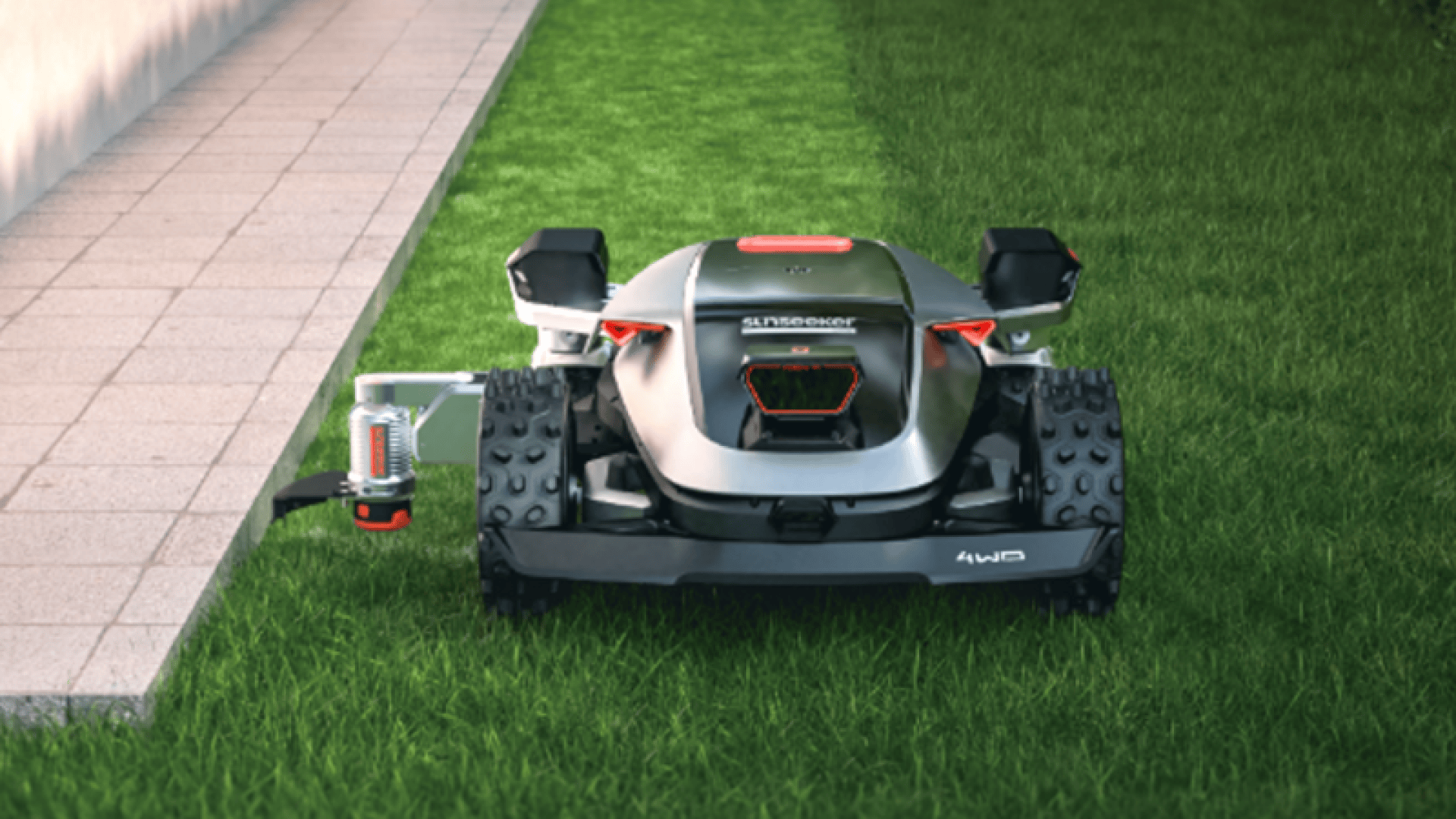 Ein Roboter-Rasenmäher mit robusten Rädern und einem Metallgehäuse mäht Gras neben einem gepflasterten Gehweg. Der Mäher ist mit einem Allradantrieb und einem sichtbaren Sensor oder einer Antenne auf der linken Seite ausgestattet.