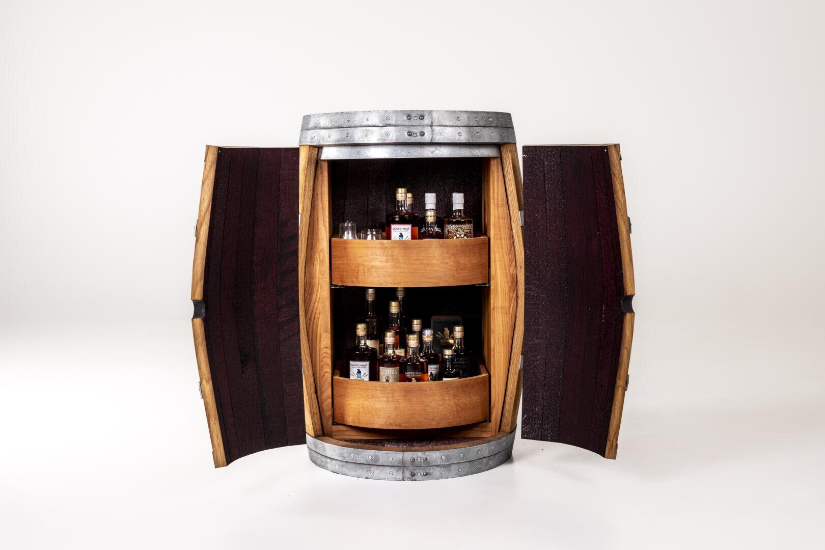 A wooden barrel with metal bands is cut open vertically to create two doors containing shelves with various bottles of schnapps. The background is plain white.