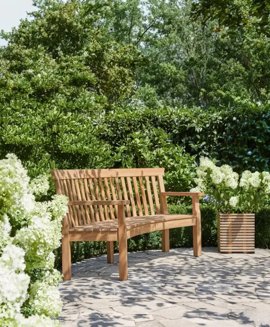 A wooden bench sits on a stone patio surrounded by lush green bushes and blooming white hydrangeas on a sunny day.