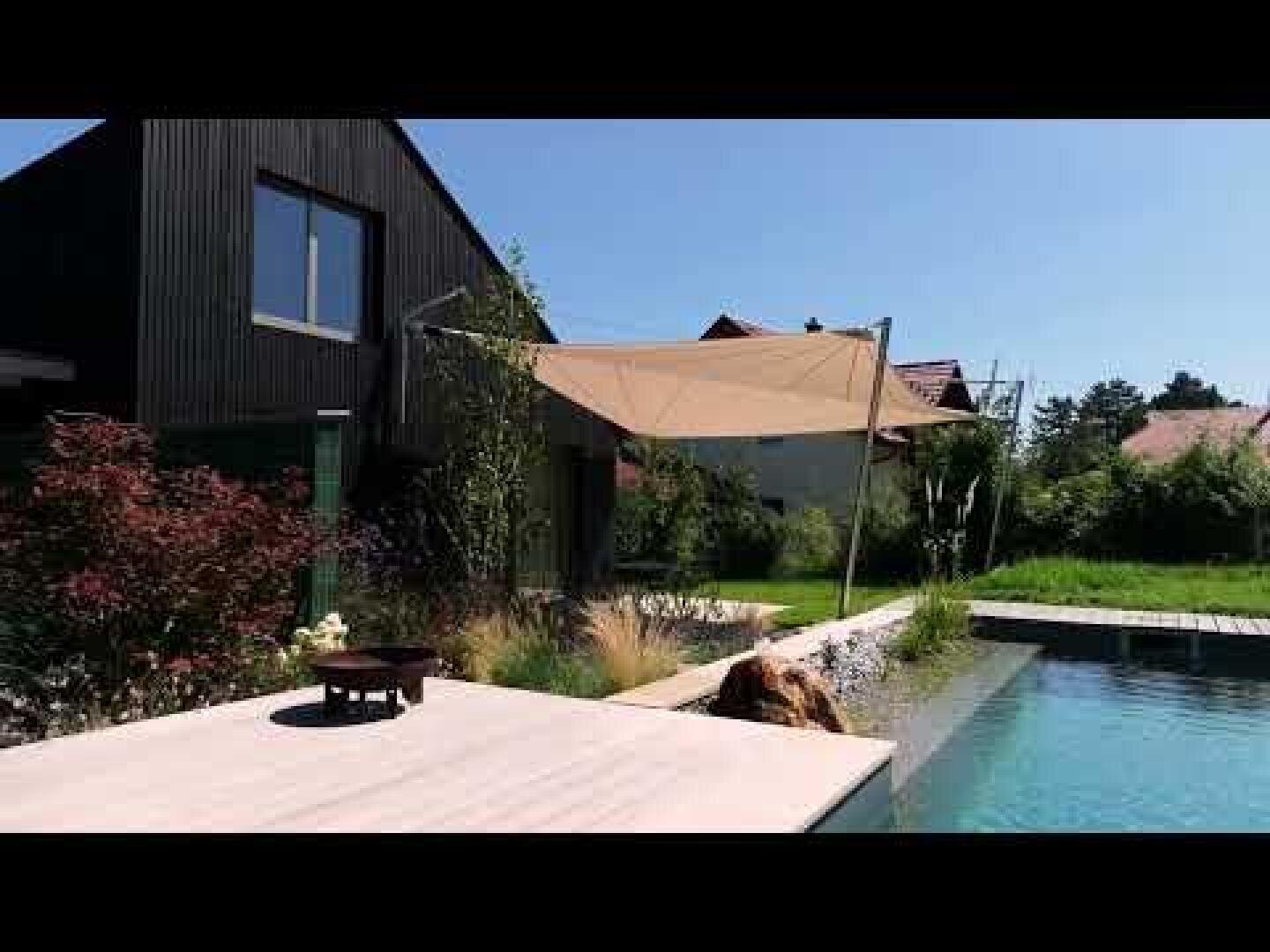 A modern, dark house with a beige awning over a terrace next to a swimming pool, surrounded by greenery and a landscaped garden on a sunny day.