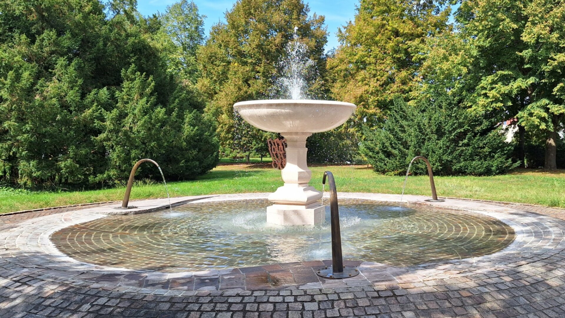 Ein runder Steinbrunnen mit einer großen zentralen Schale und drei geschwungenen Wasserspeiern aus Metall steht in einem gepflasterten Innenhof, umgeben von grünen Bäumen und Gras unter einem klaren blauen Himmel.
