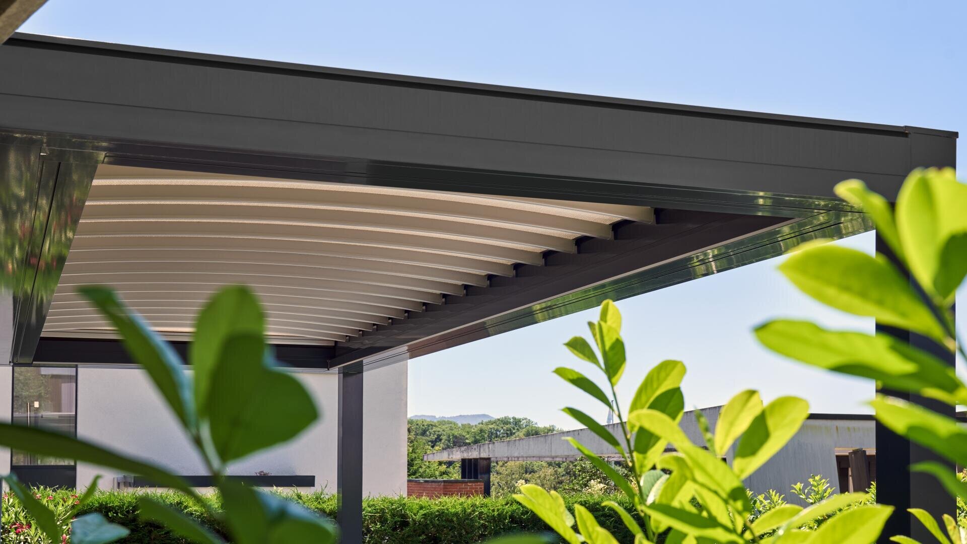A modern pergola with a slim, dark metal frame and a white slatted roof is pictured on a sunny day, surrounded by green plants and leaves, with a blue sky and buildings in the background.