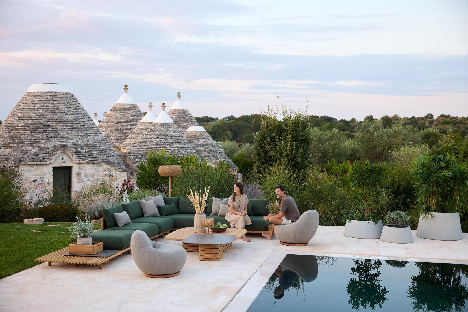 Ein Paar sitzt auf modernen Gartenmöbeln an einem Pool, umgeben von üppigem Grün und einzigartigen Trulli-Häusern mit Steindächern im Hintergrund, unter einem teilweise bewölkten Himmel.