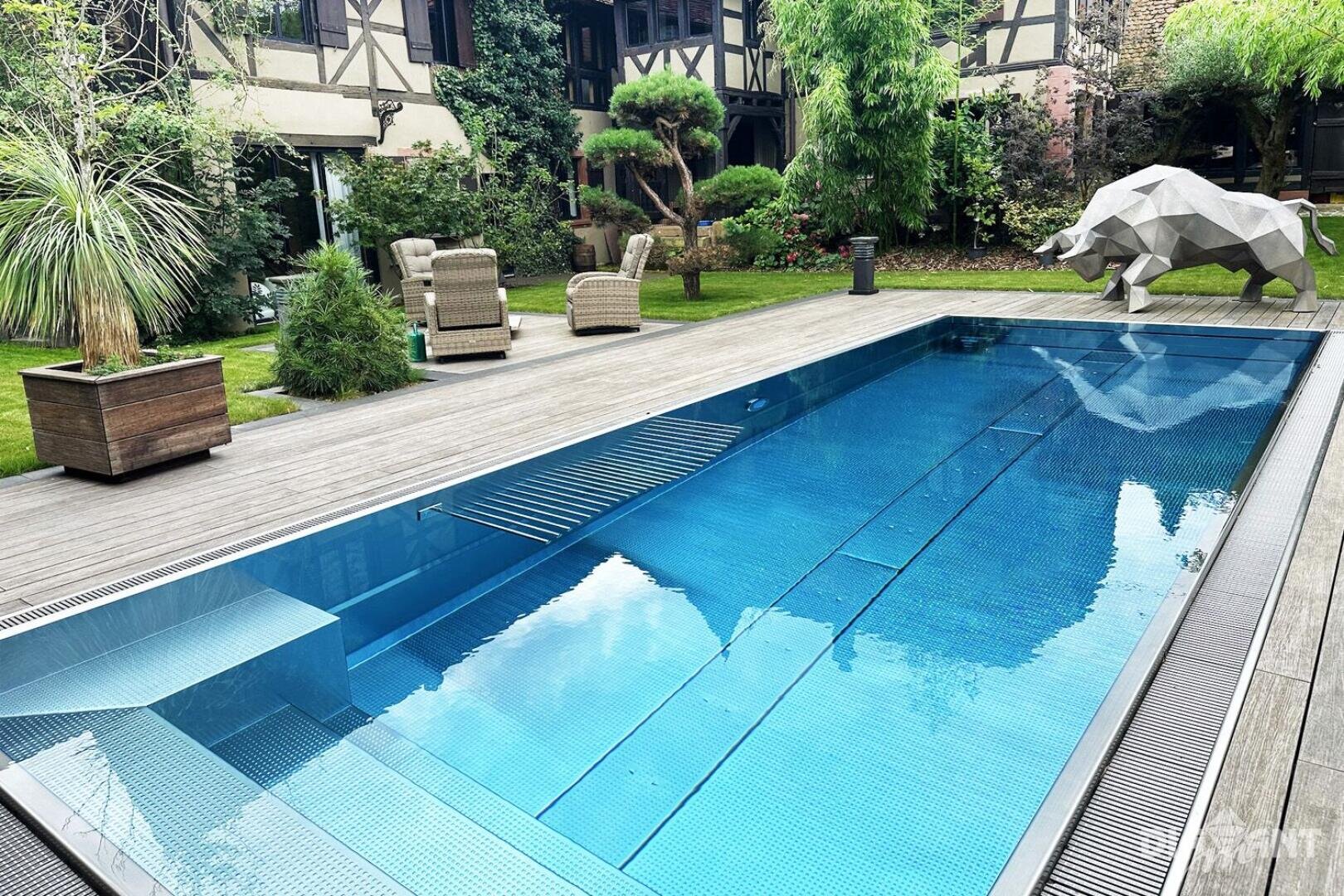 Moderner Garten mit rechteckigem Pool, Holzterrasse, Sitzgelegenheiten im Freien, üppiger Bepflanzung und einer großen metallischen Bärenskulptur am Rande des Pools.