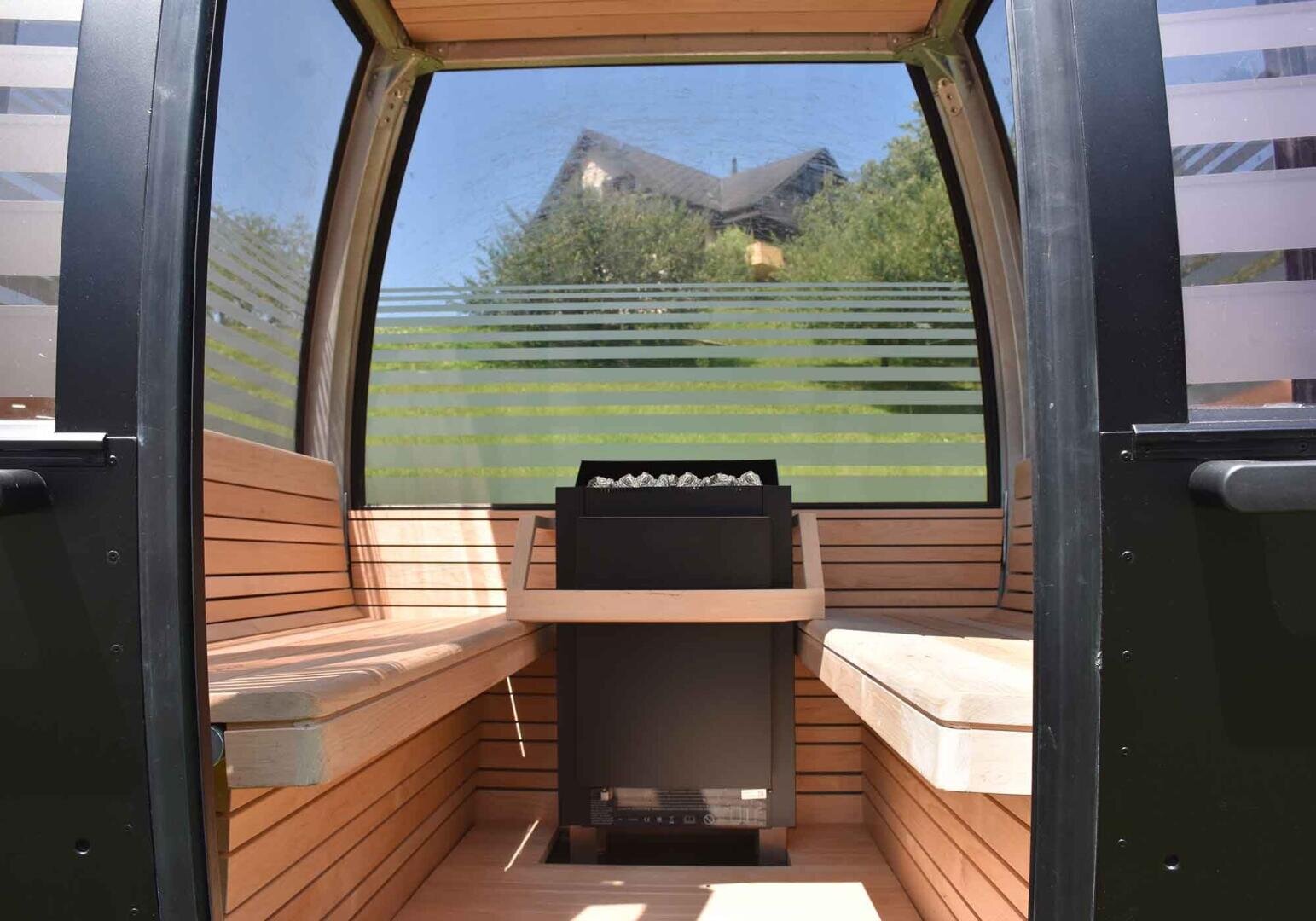 Innenraum einer kleinen, modernen Holzsauna mit Glaswänden und Bänken auf beiden Seiten. In der Mitte steht ein Ofen mit Steinen, und durch das Glas sind draußen Bäume und ein Haus zu sehen.