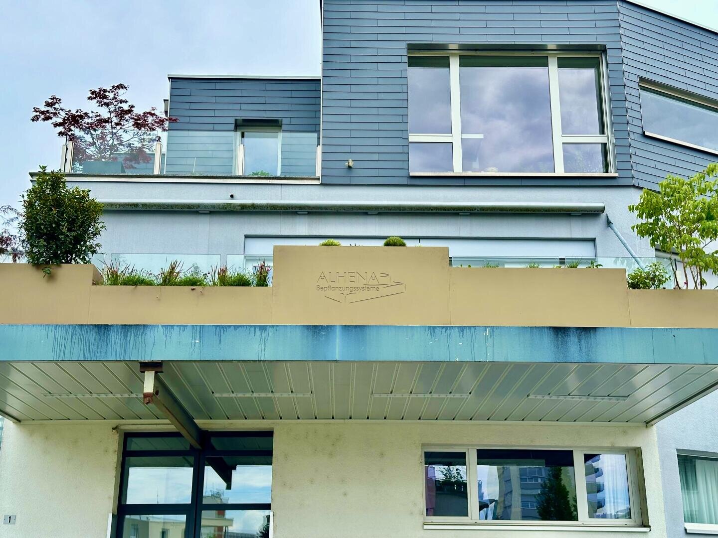 Ein modernes blaugraues Gebäude mit großen Fenstern, einem Balkon mit Pflanzen und einem beigen Schild mit der Aufschrift "ALTENAP" über dem Eingang. Der Himmel ist bewölkt, und auf dem Balkon und in der Nähe des Gebäudes ist Grün zu sehen.