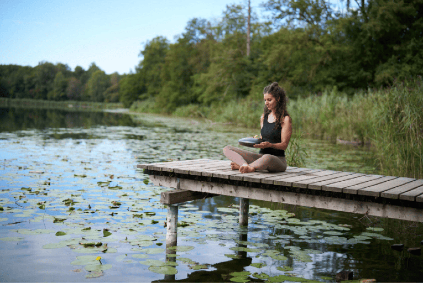 A woman sits cross-legged on a wooden dock by a lake, surrounded by lily pads and greenery, playing a handpan drum. The scene is calm and peaceful under a blue sky.