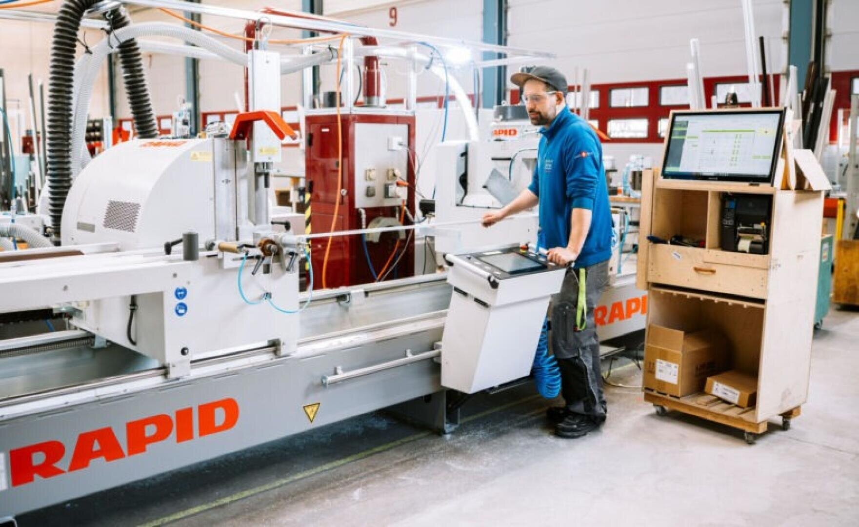 Ein Mann bedient eine große Industriemaschine mit der Aufschrift RAPID in einer Fabrik. Er steht an einem Steuerpult, und ein Computermonitor auf einem Holzwagen zeigt in der Nähe technische Daten an.