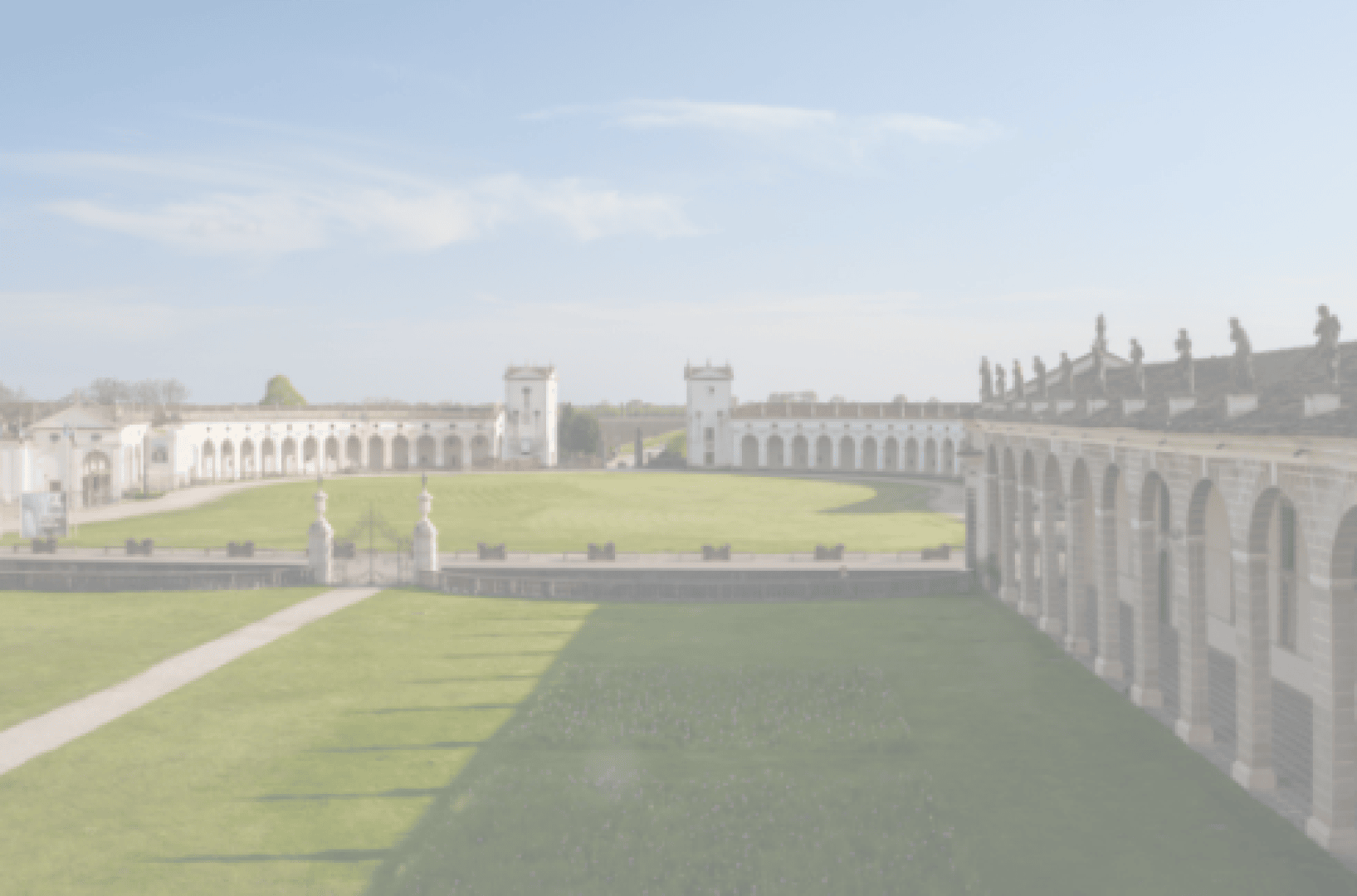 Ein großes, geschwungenes historisches Gebäude umgibt einen kreisförmigen grünen Rasen mit Türmen und Statuen. Der Himmel ist klar und blau.