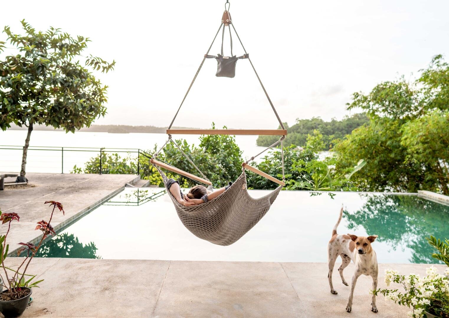 Eine Person entspannt sich in einer Hängematte an einem Infinity-Pool, mit Blick auf üppiges Grün und Wasser. Ein Hund steht auf der Veranda in der Nähe, ein weiteres Tier ist teilweise zu sehen. Die Szene ist friedlich und im Freien.