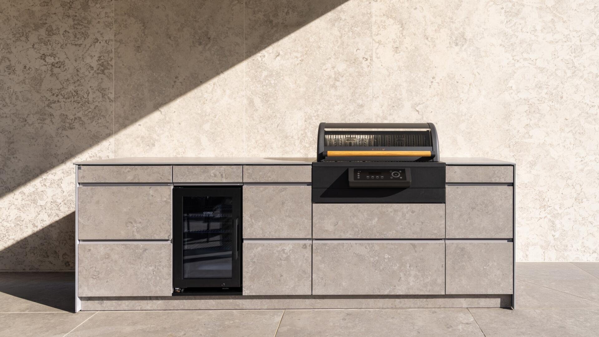 Eine moderne Außenküche aus Stein mit eingebautem Minikühlschrank und schwarzem Grill mit geöffnetem Deckel vor einer sonnenbeschienenen Steinwand.