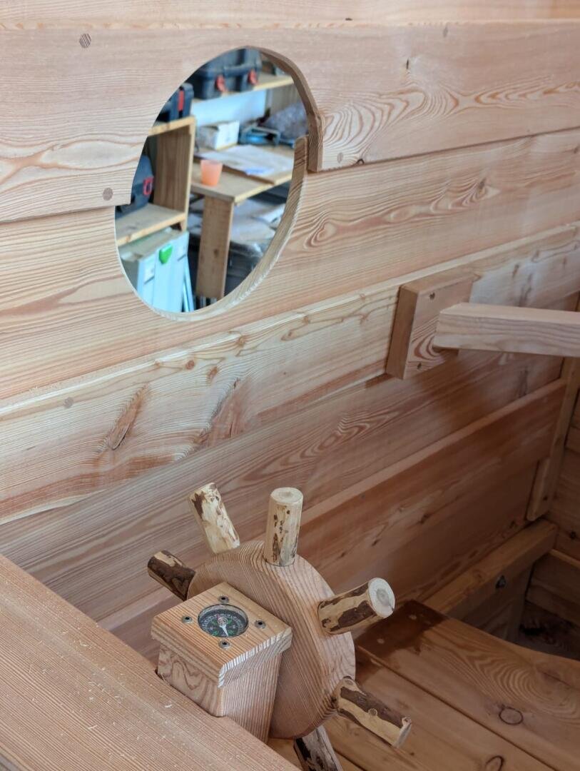 Nahaufnahme eines hölzernen Schiffsrads und Kompasses in einem handgefertigten Holzboot, mit einem runden Fenster, das eine Werkstatt im Hintergrund zeigt.