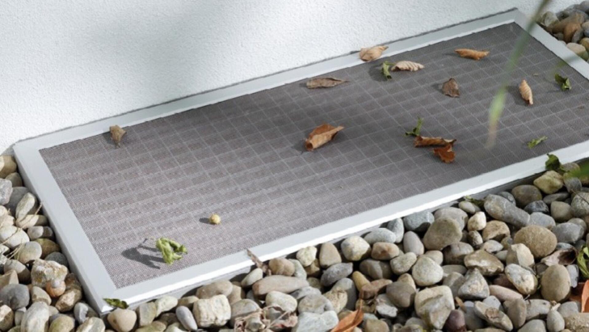 Ein Metallgitter, das einen Fensterschacht abdeckt, ist von kleinen Steinen umgeben. Oben auf dem Gitter liegen einige trockene Blätter und kleine Schuttstücke.