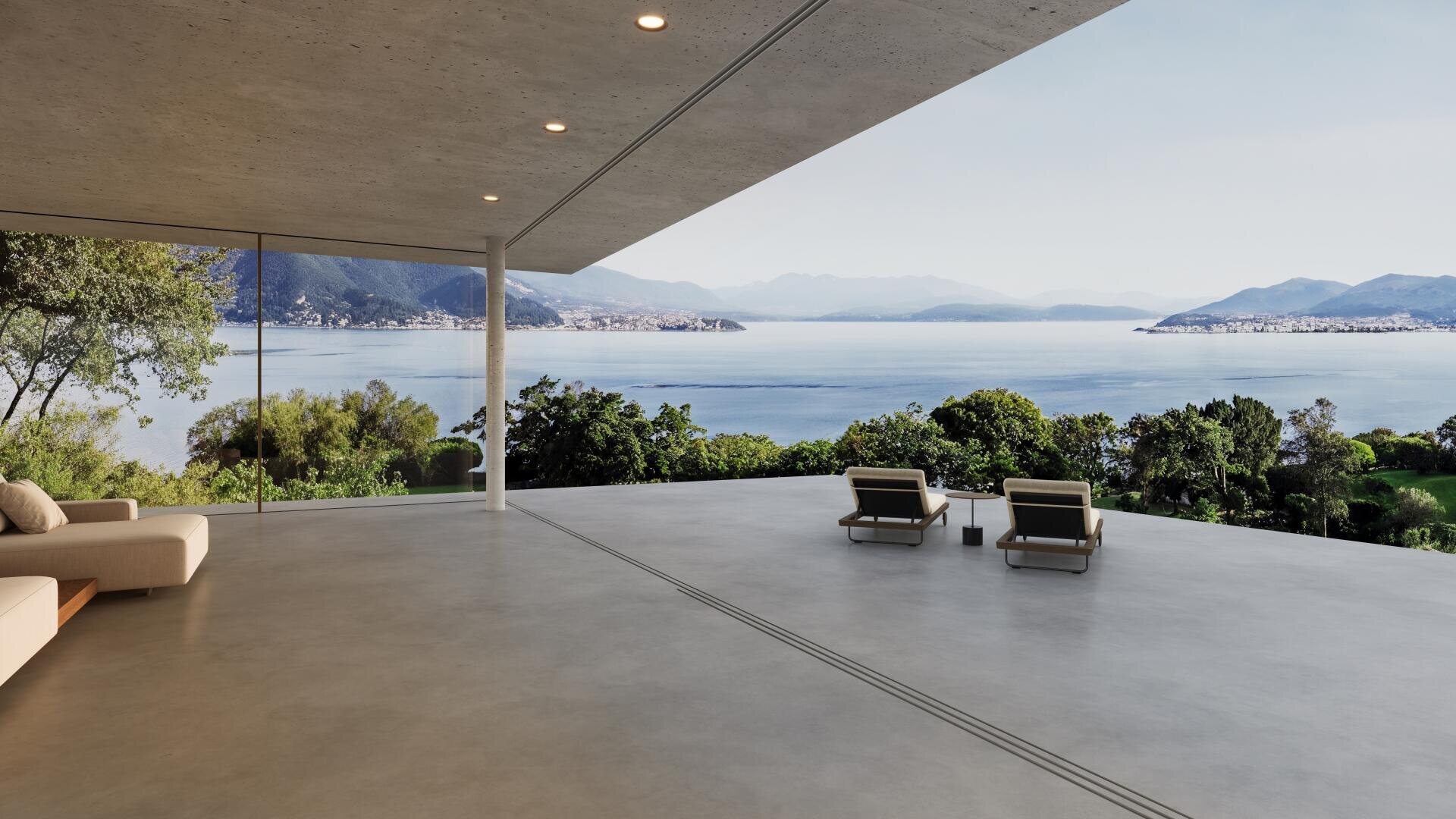 A spacious, modern terrace with two lounge chairs overlooks a picturesque lake and the distant mountains, framed by greenery and large glass walls, under a clear sky.