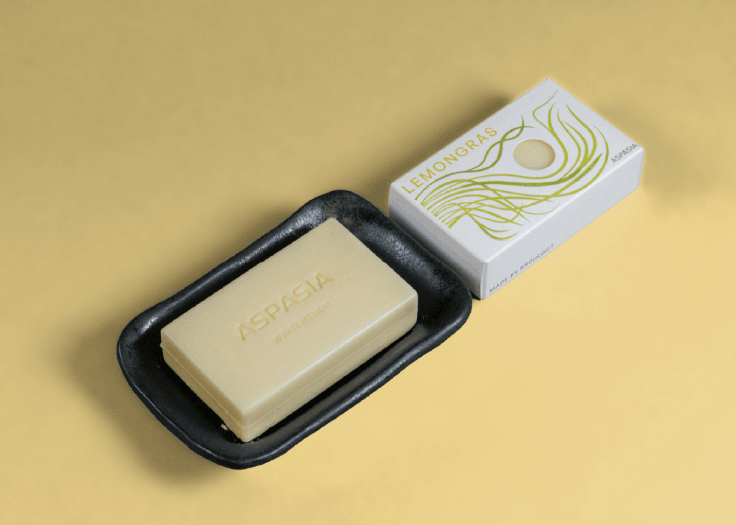 A rectangular bar of soap with the inscription ASPASIA rests on a black soap dish next to the white box with the inscription LEMONGRAS and green illustrations on a light yellow background.