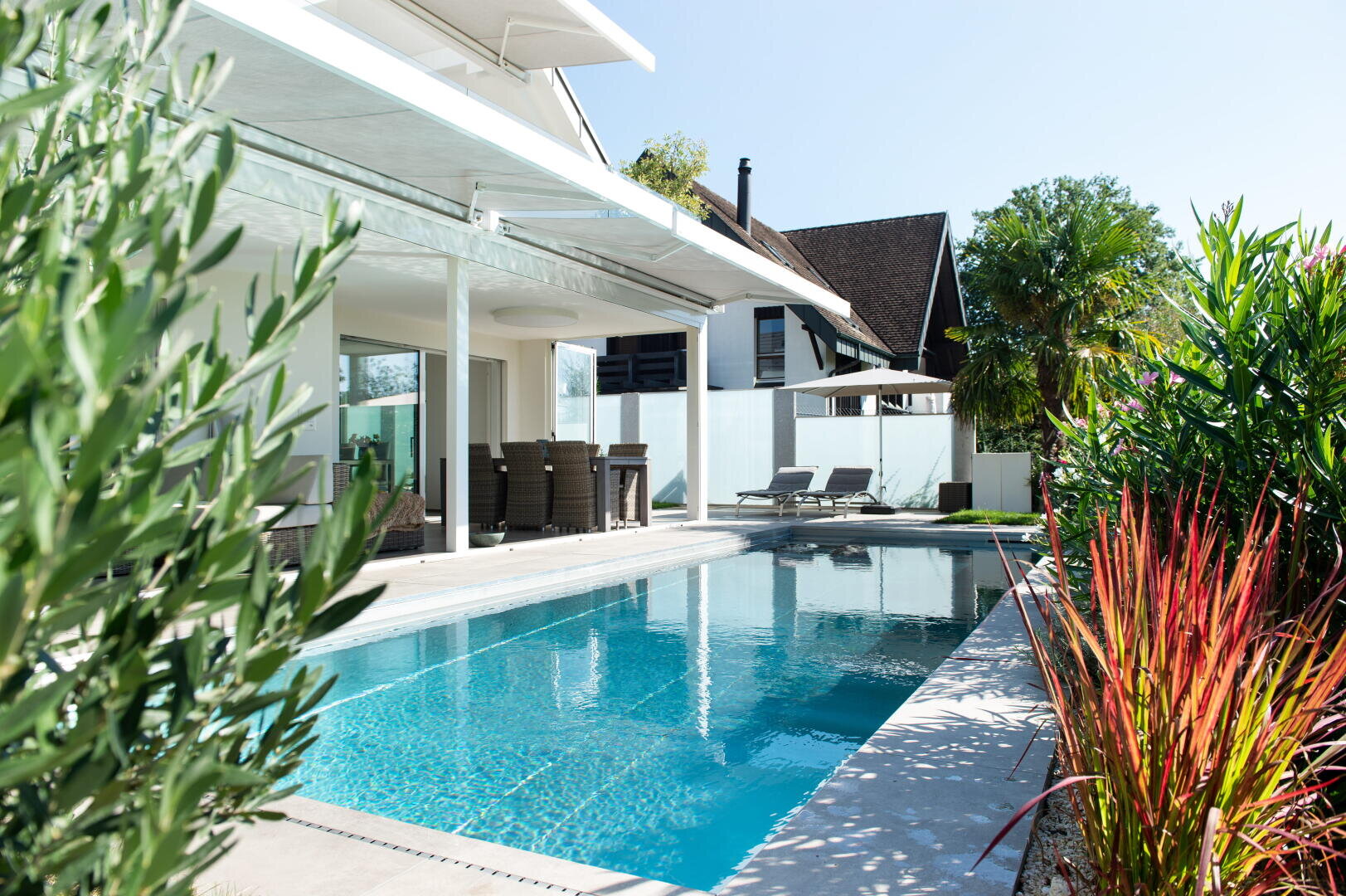 Ein modernes Haus mit einer überdachten Terrasse mit Blick auf einen rechteckigen Außenpool, umgeben von tropischen Pflanzen und Liegestühlen unter einem Sonnenschirm an einem sonnigen Tag.