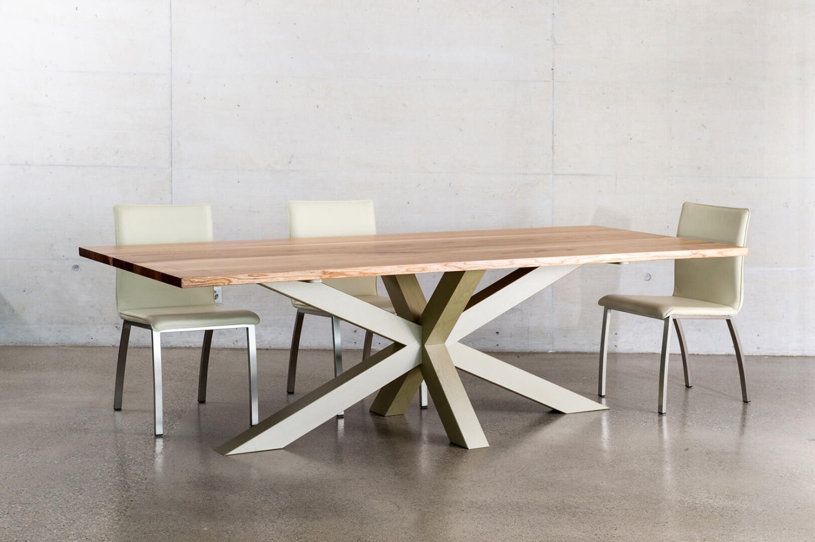 Ein moderner Esstisch aus Holz mit einem einzigartigen, geometrischen Metallgestell wird von drei hellbeigen Stühlen umgeben, die vor einer schlichten Betonwand und einem polierten Betonboden stehen.