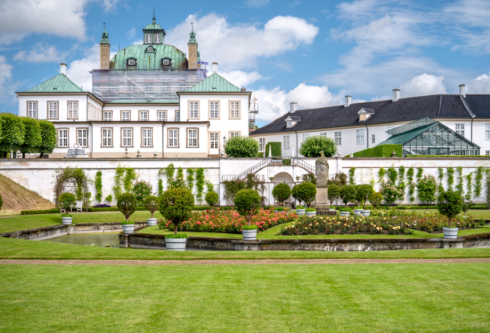 Ein historisches Herrenhaus mit grünen Dächern steht hinter gepflegten Gärten, gestutzten Hecken, runden Blumenbeeten, einem kleinen Teich und ordentlich geschnittenen Rasenflächen unter einem blauen Himmel mit Wolken.