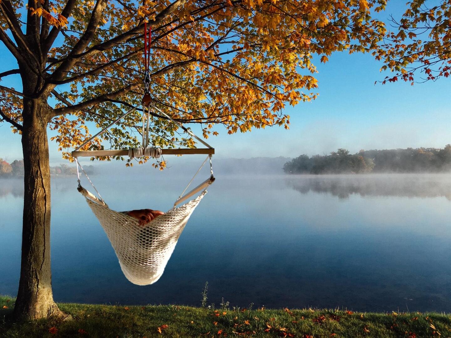 Eine Person entspannt sich in einer weißen Hängematte, die an einem Baum mit orangefarbenen Herbstblättern hängt, an einem ruhigen See mit aufsteigendem Nebel und Bäumen in der Ferne unter einem klaren blauen Himmel.